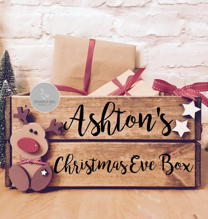Puede incluir: Una caja de madera rectangular con acabado marr&oacute;n oscuro que presenta el texto negro Ashton's Christmas Eve Box. El frente est&aacute; decorado con una figura de reno de madera con nariz roja y dos peque&ntilde;as estrellas blancas en el lado derecho.