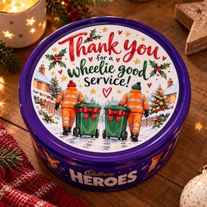 Puede incluir: Una lata redonda morada de Cadbury Heroes con una ilustración festiva de dos trabajadores con uniformes naranjas de alta visibilidad empujando contenedores verdes. El texto dice Thank you for a wheelie good service! con acebo.