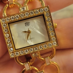 May include: A gold-coloured quartz watch with a rectangular face. The watch face is white with gold hands and the word "QUARTZ" printed at the bottom. The watch is encrusted with clear crystals around the face and on the band.