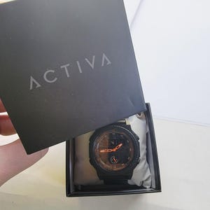 以下が含まれることがあります： 黒いACTIVAスマートウォッチ。オレンジ色のディスプレイが黒い箱に入っています。時計は丸い文字盤と黒いバンドが付いています。箱は開いており、白いクッションの上に置かれた時計が見えます。