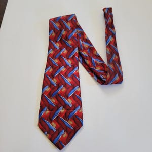 May include: A red tie with a repeating pattern of blue and gold brushstrokes. The tie is made of a smooth fabric and is displayed on a white surface. The tie's design features a diagonal pattern.
