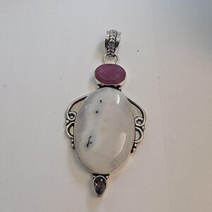 May include: A silver pendant featuring an oval, white gemstone with dark inclusions, accented by a round, pink gemstone at the top and a smaller, purple gemstone at the bottom. The pendant has decorative silver filigree.