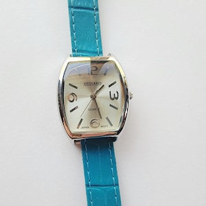 May include: A silver-toned watch with a rectangular face and a teal blue leather band. The watch face displays the brand name "KESSARIS" and the numbers 12, 3, and 6. The watch is a quartz movement.