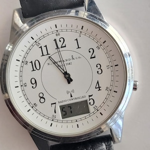 Könnte beinhalten: Eine silberfarbene Armbanduhr mit weißem Zifferblatt, schwarzen Ziffern und Zeigern. Das Zifferblatt zeigt den Text "E. Howard & Co. 1842" und "Radio Controlled". Die Digitalanzeige zeigt "57". Das Armband ist aus schwarzem Leder.
