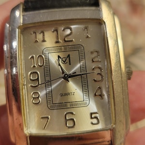May include: A rectangular quartz watch with a silver-colored face and gold-colored frame. The watch face displays black numerals and hands, with the word "QUARTZ" at the bottom. The watch has a black leather band.
