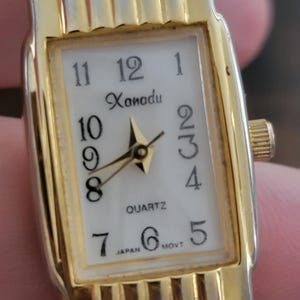 Könnte beinhalten: Eine rechteckige Armbanduhr in Goldoptik mit weißem Zifferblatt. Das Zifferblatt zeigt das Wort "Xanadu" über den Zeigern, mit schwarzen Ziffern und goldenen Zeigern. Die Uhr ist mit "QUARTZ" und "JAPAN MOVT" gekennzeichnet.