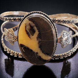Puede incluir: Brazalete de puño plateado con una piedra preciosa ovalada marrón y amarilla. La piedra preciosa está engastada en un bisel plateado con un diseño de cuerda trenzada. El brazalete tiene detalles florales y bandas decorativas.