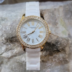 May include: A white Anne Klein New York watch with a gold-tone bezel encrusted with crystals. The watch face is white with gold Roman numerals and hands. The watch band is white.