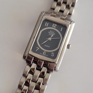 May include: A rectangular silver wristwatch with a dark grey face. The watch face displays the brand "Bill Robin Son" and the word "Quartz". The watch has a silver link band.