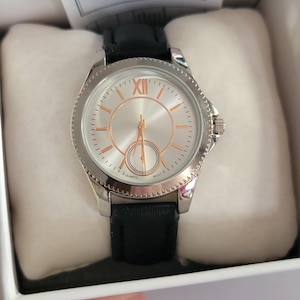 Könnte beinhalten: Silberfarbene Quarzuhr mit schwarzem Lederarmband. Das Zifferblatt der Uhr ist silberfarben mit roségoldfarbenen Zeigern und römischen Ziffern. Die Uhr befindet sich in einer weißen Box.