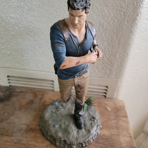 Puede incluir: Figura detallada de un hombre con camisa azul, pantalones color caqui y botas, de pie sobre una base rocosa. La figura tiene una expresión seria y sostiene un objeto pequeño. La estatua tiene correas de cuero marrón y detalles visibles como manchas de sangre en el brazo.