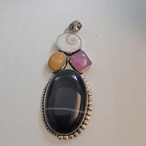 May include: A silver pendant featuring an oval black stone with a white stripe, surrounded by a beaded border. Above it are a white spiral, a yellow oval, and a pink square stone. The pendant has a decorative bail.