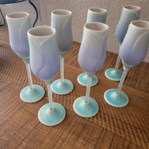 May include: Set of seven tulip-shaped champagne flutes. The glasses feature a gradient of colors, transitioning from white to light blue and lavender. Each glass has a white stem and a light blue base, creating an elegant aesthetic.