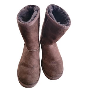 May include: A pair of mid-calf boots in a dark brown suede finish. The boots feature a rounded toe, a flat sole, and a thick, dark brown plush lining visible at the top opening. The exterior surface has a soft, matte texture with visible stitching.