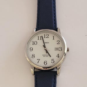 Könnte beinhalten: Eine Timex Armbanduhr mit rundem silberfarbenem Gehäuse, weißem Zifferblatt mit schwarzen arabischen Ziffern und einem Datumsfenster auf der Drei-Uhr-Position. Die Uhr hat ein dunkelblaues Stoffarmband und zeigt den Text Indiglo und WR 30M.