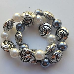 May include: A bracelet featuring a mix of beads in silver, pearl white, and dark gray. The bracelet includes round pearl beads, silver spiral-design beads, and oval silver beads with etched patterns. The beads are strung together to form a circular shape.