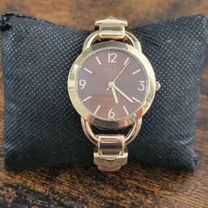Peut inclure: Une montre Studio dorée avec un cadran marron et des aiguilles et index dorés. La montre a un cadran rond et un bracelet. La montre repose sur un coussin noir texturé.