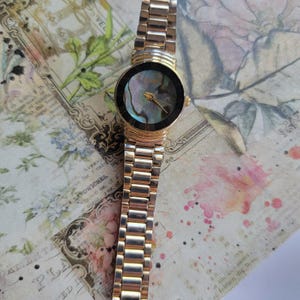 May include: A gold-toned wristwatch with a round face and a mother-of-pearl dial. The watch has a linked band and Roman numeral hour markers. The watch is displayed on a vintage paper background with floral and text elements.