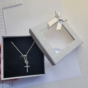 May include: A silver necklace with a cross pendant, displayed in an open, glittery silver gift box with a clear window and a silver bow. The cross pendant is adorned with small, clear stones. The box is on a white surface.
