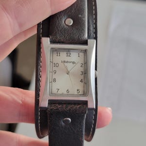 May include: A rectangular Billabong watch with a silver-tone case and a light-colored face. The watch has black numbers and hands. The watch is attached to a wide, dark brown leather band with silver rivets.