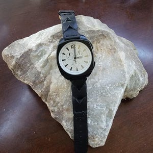 May include: A black wristwatch with a white face and black leather strap. The watch has a square face with a round dial, featuring black numerals and hands. The strap has a unique chevron design.