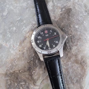May include: A silver-toned wristwatch with a black face and a black leather band. The watch face features white numerals, a red second hand, and the text "SWISS ARMY". The watch is set against a textured, gray surface.