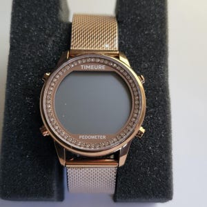 May include: Rose gold smartwatch with a mesh band. The watch face is surrounded by a ring of small, clear crystals. The watch face displays the word "TIMEURE" above the screen and "PEDOMETER" below.