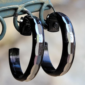 May include: A pair of black hoop earrings with a silver geometric design. The earrings have a hinged closure.