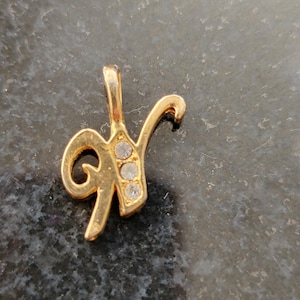 May include: Gold-colored pendant in the shape of the letter "Y". The pendant features three clear, round gemstones and a loop for a chain. The jewelry is set against a dark, textured background.