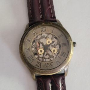 May include: A vintage-style wristwatch with a bronze-colored case and a brown leather band. The watch face features Roman numerals and a skeleton design, revealing the inner workings. The watch has the letters "AEO" on the face.