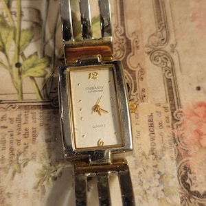 Puede incluir: Un reloj de cuarzo Embassy by Gruen rectangular con una correa de metal plateado y dorado. La esfera del reloj es blanca con manecillas doradas y los números 12 y 6. El reloj está sobre un fondo estampado.