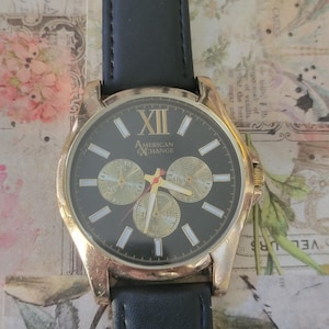 May include: A gold-toned wristwatch with a black face and a black leather band. The watch face features gold-colored hands and markers, with the text "AMERICAN EXCHANGE" and the Roman numeral XII. Three smaller dials are visible.