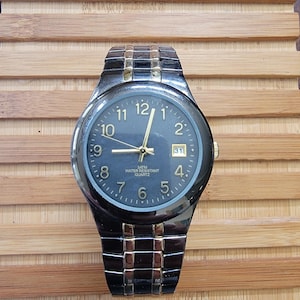 May include: A black and gold wristwatch with a dark blue face. The watch has gold hands and hour markers, a date display, and a black metal band. The watch is water-resistant and has a quartz movement.