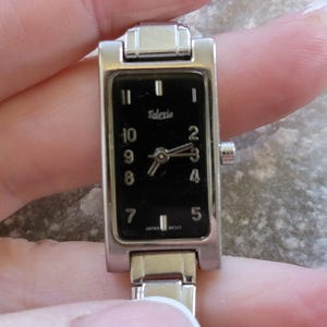 Könnte beinhalten: Eine rechteckige, silberfarbene Talexio-Armbanduhr mit schwarzem Zifferblatt. Die Uhr hat weiße Ziffern und Zeiger, und der Text "Talexio" ist über den Zeigern aufgedruckt. Das Armband der Uhr ist ebenfalls silberfarben.
