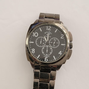 May include: A silver-toned metal wristwatch with a black circular dial featuring the text Beverly Hills Polo Club and Heritage. The watch includes three sub-dials, white hour markers, and large white numerals at the 2, 4, 6, 8, 10, and 12 positions.
