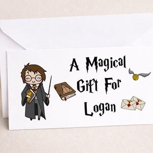 May include: White card with the text "A Magical Gift For Logan" in a stylized font. The card features a cartoon illustration of a wizard, a book, a golden snitch, and two envelopes.