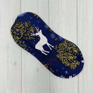 May include: A navy blue reusable menstrual pad with a white deer silhouette and gold leaf pattern. The pad features a whimsical forest design with stars and floral accents, offering an eco-friendly alternative.