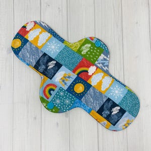 May include: A reusable cloth menstrual pad with a colourful patchwork design featuring weather-related illustrations. The pad has a blue backing and is secured with two teal press studs. The design includes rainbows, suns, clouds, and snowflakes.