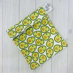 Puede incluir: Una bolsa de sándwich reutilizable blanca con un estampado de aguacate lindo. La bolsa tiene una parte superior plegable y una etiqueta que dice "Hecho con amor".