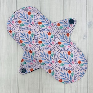 Puede incluir: Una almohadilla de tela reutilizable rosa con un patrón floral y palabras como "paz, amor, compasión, honestidad, amabilidad, crecimiento, empathie, humildad, apertura, curiosidad, felicidad, resiliencia, actitud" impresas en ella.