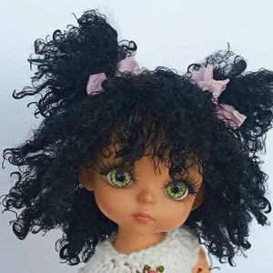 Könnte beinhalten: Eine Puppe mit lockigem schwarzem Haar, grünen Augen und einer rosa Schleife in jedem Zopf. Die Puppe trägt ein weißes Häkelkleid.