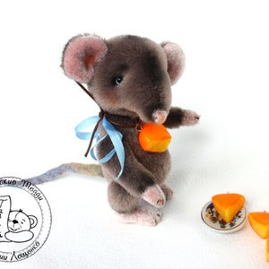 Puede incluir: Un juguete de peluche de ratón gris que sostiene un pequeño trozo de queso naranja. El ratón lleva una cinta azul alrededor del cuello. Hay dos trozos de queso naranja más en un pequeño plato blanco con un diseño floral.