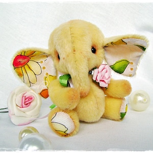 May include: A yellow plush elephant toy with a floral print on its ears. The elephant is holding a pink rose in its trunk. The toy is sitting on a white surface.
