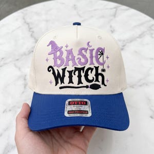 May include: A cream-colored baseball cap with a blue brim featuring the embroidered purple and black text "BASIC WITCH" with a witch hat, spider web, and broomstick graphic. A rectangular sticker on the brim reads "OTTO CLASSIC OSFM".