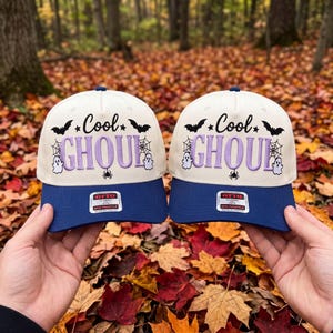 May include: Two off-white baseball caps with navy blue brims, each featuring the embroidered text "Cool GHOUL" in purple, surrounded by black graphic icons of bats, spiderwebs, ghosts, and a spider, with an Otto brand sticker on the brim.