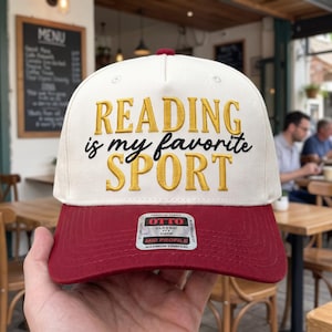 May include: A cream-colored baseball cap with a maroon brim. The front features embroidered text in gold and black that reads READING is my favorite SPORT. A rectangular sticker on the brim displays the brand name OTTO and product details.