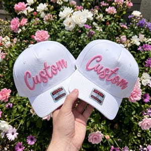 May include: Two white baseball caps featuring the word Custom embroidered in raised pink script lettering on the front panels. Each cap includes a silver and black Otto brand sticker on the brim, indicating a mid-profile classic design.