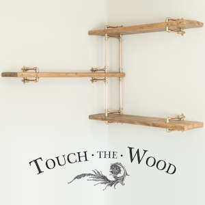 May include: Three wooden shelves with copper pipe supports mounted on a white wall. The shelves are in a corner and the top shelf is the longest. The text "TOUCH. THE. WOOD" is on the wall below the shelves.