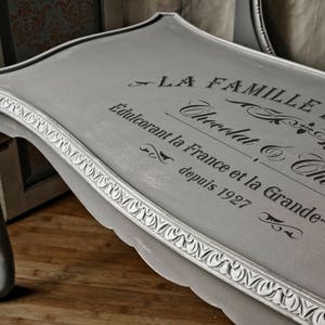 May include: A gray painted table with ornate detailing and a black stencil design that reads "La Famille de Petits Chocolat & Churros Edulcorant la France et la Grande-Bretagne depuis 1927".