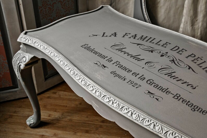 May include: A gray painted table with ornate detailing and a black stencil design that reads "La Famille de Petits Chocolat & Churros Edulcorant la France et la Grande-Bretagne depuis 1927".